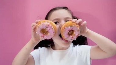 Cute kid holding donuts with icing in her hands and trying to it them, makes glasses. Pink background.
