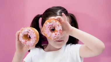 Cute girl holds sweet donuts with icing in her hands, smiling, looking at the camera and makes glasses. Pink background