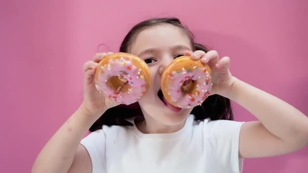 Cute kid holding donuts with icing in her hands and trying to it them, makes glasses. Pink background. — Stockvideo