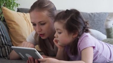 Ciddi anne-kız evdeki kanepede dijital tablet kullanıyor. Çocuk teknolojiyi sosyal uzaklık ve kendini izolasyon sırasında Coronavirus karantinasında öğreniyor. Kapat.