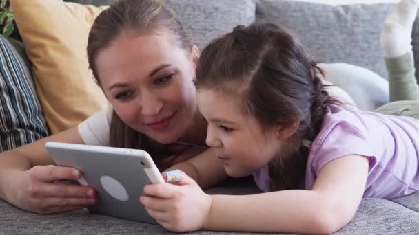 Ibu yang bahagia dan putri kecilnya menggunakan tablet digital di sofa di rumah. Anak perempuan belajar teknologi selama jarak sosial dan isolasi diri dalam penguncian karantina — Stok Video
