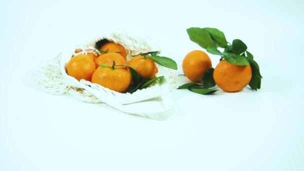 Мандарины катятся на белый многоразовый пищевой мешок с другими мандаринами на столе с белым фоном. Концепция переработки отходов — стоковое видео