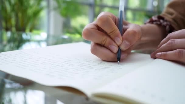 Femme écrit sur un bloc-notes en papier ouvert avec stylo à bille noir assis à une table en verre dans un café avec des plantes autour. Gros plan — Video