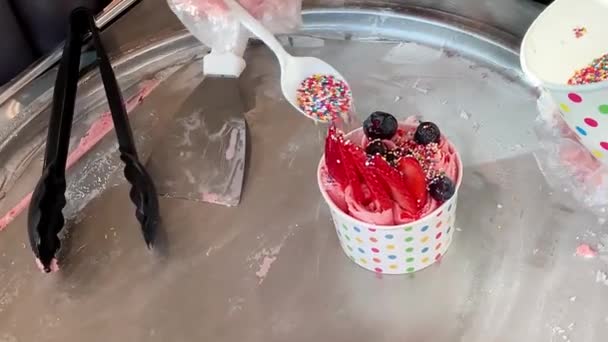 A nő színes öntözéssel díszíti a kevert sült eper fagylalt tekercseket egy papírpohárban. Házi készítésű édes desszert — Stock videók