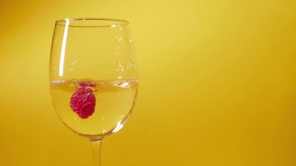 De cerca. Tres frambuesas cayendo en copa de vino blanco sobre fondo amarillo. Burbujas alrededor de las bayas — Vídeos de Stock