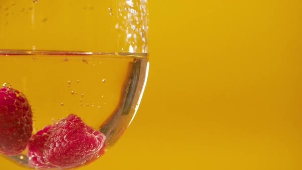 Makroextreme Nahaufnahme. Himbeere fällt in ein Glas Weißweinalkohol vor gelbem Hintergrund. Blasen um Beeren — Stockvideo