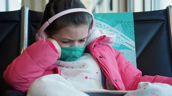 Yeşil koruyucu maskeli ve pembe kulaklıklı sıkılmış çocuk havaalanında tabletle çizgi film izliyor. Yeni gerçeklik kavramı