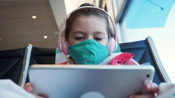 Kahverengi saçlı, yeşil koruyucu maskeli ve havaalanında tablet kullanan pembe kulaklıklı anaokulu öğrencisi kız. Yeni gerçeklik kavramı