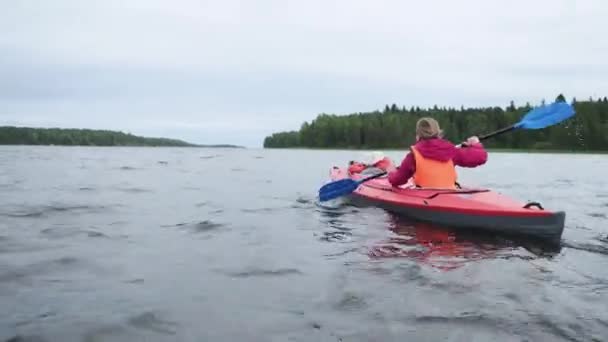 Wanita muda berkacamata mendayung kayak olahraga oranye terang dengan anak kecil berlayar di sepanjang danau melawan hutan dan langit abu-abu mendung pada hari yang buruk — Stok Video