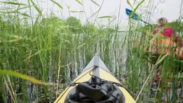 Unrecognizable mother with kid does kayaking by yellow plastic sports boat across large green thickets of reeds under cloudy sky on summer. View from nose of ship — Stock Video