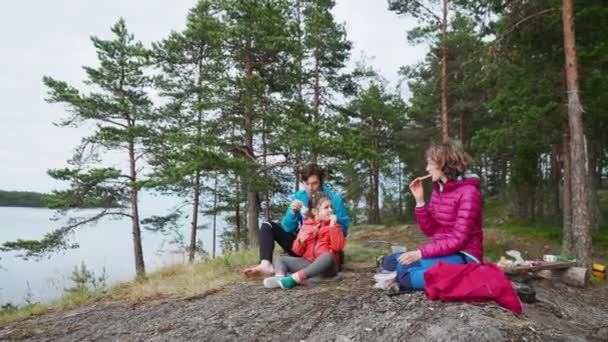 4K Счастливая молодая семья на весеннем кемпинге на озере. Два родителя вместе с красивой дочерью сидят перед озером и едят. Семейные отношения Деятельность — стоковое видео