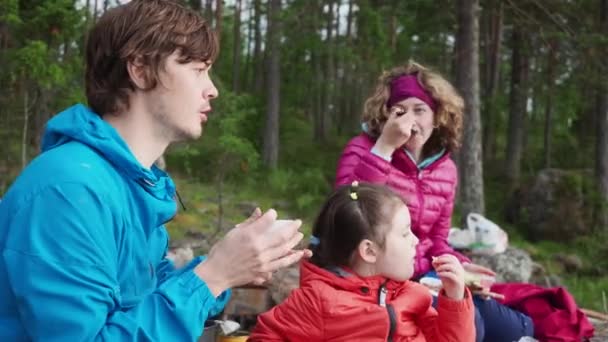 Aktivität in Familienbeziehungen. Glückliche junge Familie im Frühling Campingurlaub, Kiefernwald Hintergrund. Zwei Eltern unterhalten sich beim Frühstück, hübsche Tochter sitzt nachdenklich in der Nähe und isst. — Stockvideo