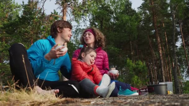 4K Två lyckliga föräldrar kysser varandra på vårcampingen medan stygga dottern visar tungan. Vår camping familjesemester vid sjön nära tallskog bakgrund. — Stockvideo