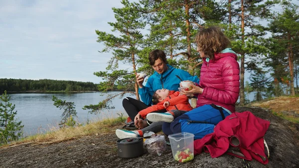 4K Familia feliz de dos padres jóvenes y chica bastante juguetona en vacaciones de campamento de primavera en el lago cerca de bosque de pinos de fondo comiendo el desayuno de vajilla reutilizable —  Fotos de Stock