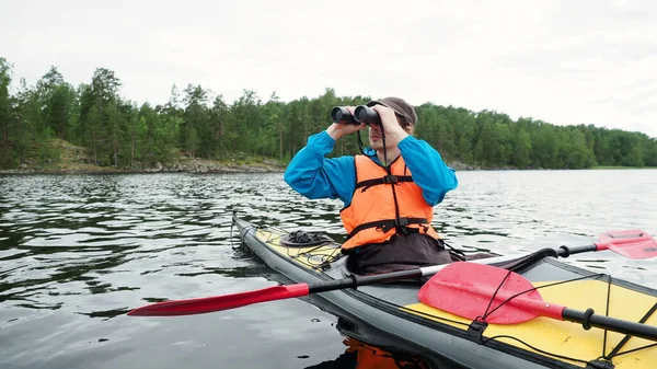 El hombre preocupado mira a través de prismáticos sentados en kayak amarillo gris en el día de verano. Actividades al aire libre, concepto de deporte acuático. —  Fotos de Stock