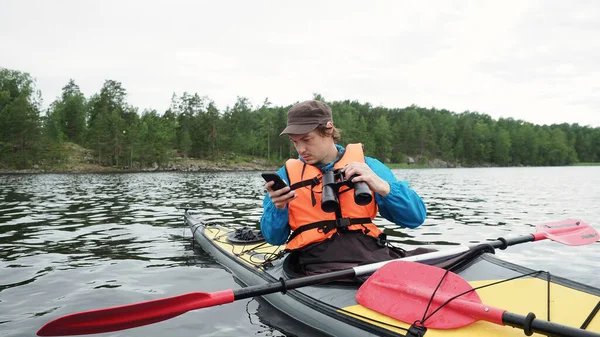 Aggódó ember online térkép ellenőrzése mobil eszköz kezében távcső és ül szürke sárga kajak hajó a nyári napon. Szabadtéri tevékenységek, vízi sportok koncepciója. — Stock Fotó