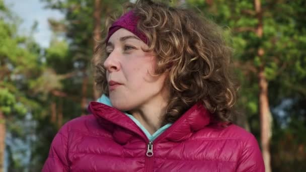 De cerca. Feliz cabello rizado mujer joven comiendo y sonriendo misteriosamente en vacaciones de campamento de primavera, fondo de bosque de pinos. Concepto de naturaleza trekking familiar. — Vídeo de stock