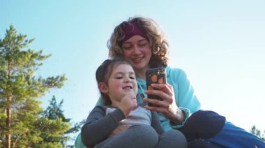 Kapatın. Mutlu kıvırcık saçlı anne ve küçük kızı sohbet ediyor. Bahar kampı tatilinde gün batımında cep telefonu uygulamalarıyla servis ağı oynuyor. Doğanın nitelikli aile zamanı kavramı