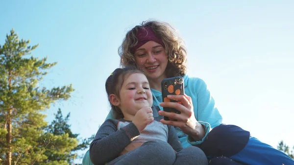 Kapatın. Mutlu kıvırcık saçlı anne ve küçük kızı sohbet ediyor. Bahar kampı tatilinde gün batımında cep telefonu uygulamalarıyla servis ağı oynuyor. Doğanın nitelikli aile zamanı kavramı