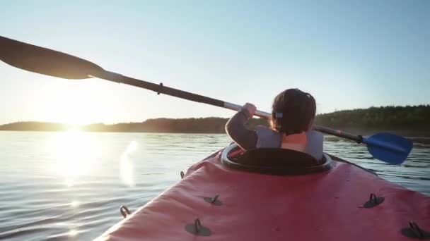 Kindergartener child learning to sail small red kayak along large tranquil lake against dark forest at bright sunset in summer evening — Stock Video