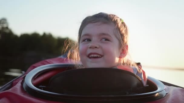 Schattige kleuter meisje blij glimlachen naar de camera zitten in de kleine rode kajak langs rustige meer tegen donker bos bij heldere zonsondergang in de zomer avond — Stockvideo