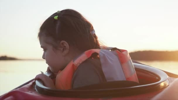 Verontrustend en verontrustend jong meisje kind kijkt in het water zitten in de kleine rode kajak langs rustige meer tegen donker bos bij heldere zonsondergang in de zomer avond — Stockvideo