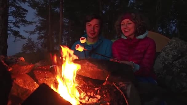 Familia feliz de turistas pareja en un viaje. joven hombre y mujer asan malvaviscos dulces en hoguera de campamento con esquiadores de metal. El concepto de estilo de vida familiar en vacaciones. Movimiento lento — Vídeo de stock