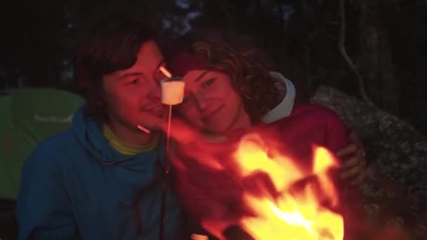 閉めろ。笑顔の観光客の旅にカップル。キャンプのたき火で金属製の串焼きに甘いマシュマロをローストしながら抱きつく若い男と女。休日の家族のライフスタイルの概念。スローモーション — ストック動画