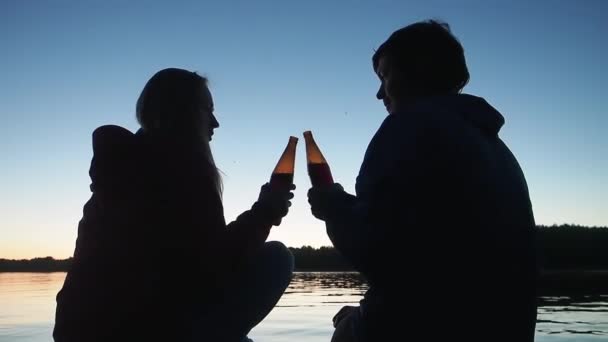 Siluetas de pareja joven tintineando botellas oscuras de deliciosa cerveza sentada cerca de un gran río tranquilo al atardecer vista trasera — Vídeo de stock