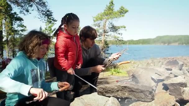 Pareja joven casada con una hija pequeña descansando en la naturaleza, asando salchichas a fuego alto cerca del lago en el bosque, picnic familiar el fin de semana. Acampar en vacaciones de verano. Estilo de vida itinerante — Vídeos de Stock