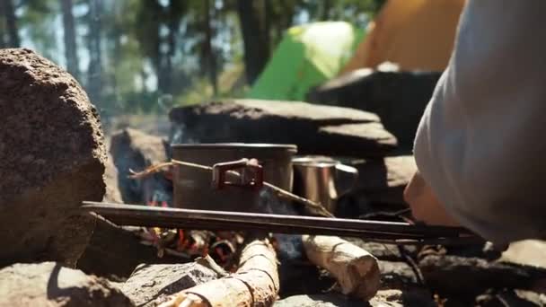 Resenär matlagning i slev på elden. Okänd hand sätter en kastrull på en sten nära elden på bakgrunden nära skogen. Familjepicknick på helgen. Camping på sommarsemester. Resande livsstil — Stockvideo