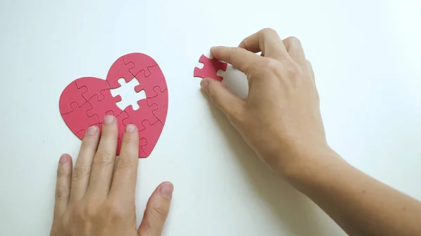 Kadınların elleri, kırmızı kalp şeklindeki yapbozun kayıp parçalarından birini beyaz masaya koyar. Üst görünüm