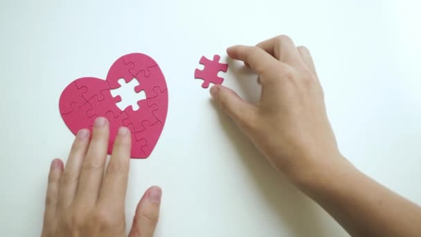 Vrouwen handen plaatst een stukje puzzel op het ontbrekende deel om rode hartvormige decoupeerzaag op witte tafel. Bovenaanzicht — Stockvideo