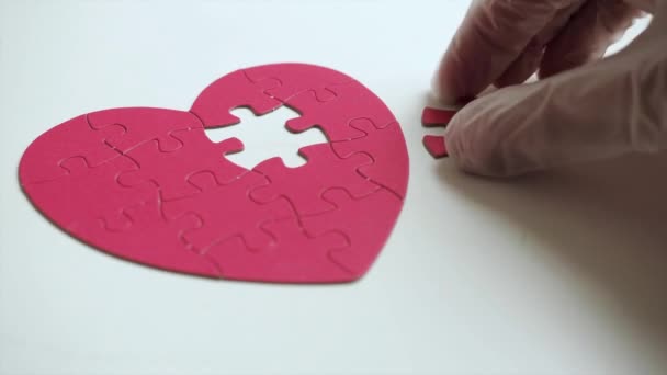 Extrém közelről. Nők kezét fehér orvosi latex kesztyű helyezi egy darab puzzle a hiányzó rész piros szív alakú kirakós fehér asztalon. Felülnézet — Stock videók