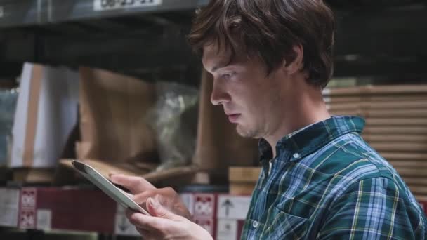 Extreme close up. Concentrated young brunette guy employee in a plaid shirt with tablet checking goods in cardboard packages standing on high shelves in spacious warehouse — Stock Video