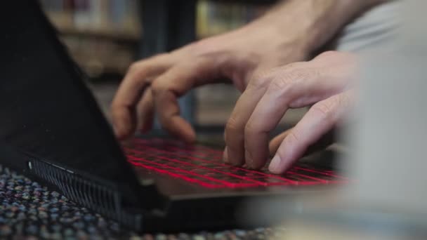 Közelről. Felismerhetetlen emberek kezek gépelés gyorsan egy laptop fekete billentyűzet piros háttérvilágítással a könyvtárban, könyvespolcok háttér. Férfi vállalkozó a könyvesboltban vagy az irodában — Stock videók