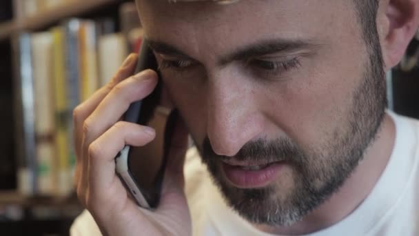 Kapatın. Beyaz tişörtlü, orta yaşlı, sakallı bir adam kütüphane kitaplıklarında telefonla ciddi ciddi konuşuyor. Erkek girişimci ya kitapçıdadır ya da işyerindedir. Kapat. — Stok video