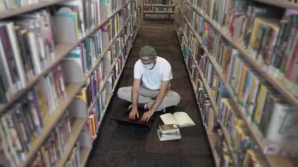 Pria paruh baya yang serius dengan topeng medis pelindung dengan kaos putih mengetik dengan cepat pada papan ketik hitam laptop di perpustakaan, latar belakang rak buku. Kehidupan baru setelah pandemi. Tutup. — Stok Video