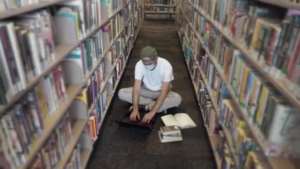 Grave uomo di mezza età con maschera medica protettiva in bianco T-shirt digitando rapidamente sulla tastiera nera portatile in biblioteca, scaffali sfondo. Nuova vita dopo la pandemia. Da vicino. — Video Stock