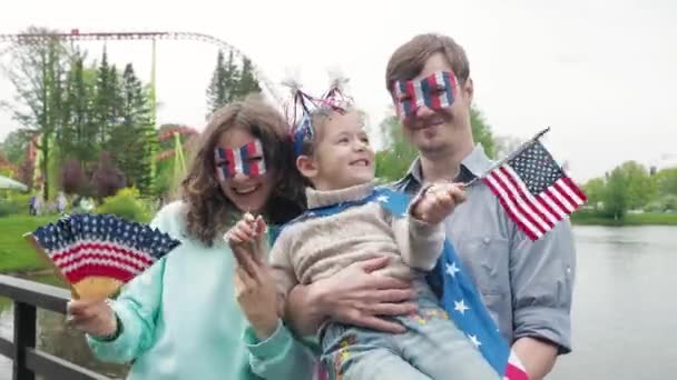 Vooraanzicht. Vier het op 4 juli. Portret van een gelukkig jong gezin buiten in een park tijdens Onafhankelijkheidsdag, kijkend naar de camera, zwaaiend met Amerikaanse vlaggen. — Stockvideo