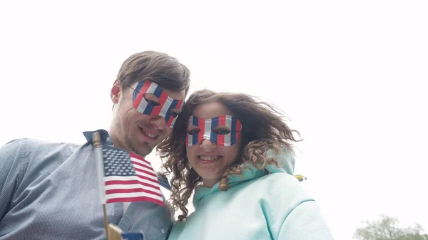 4 Temmuz Bağımsızlık Günü 'nde parkta dikilen mutlu genç çift mavi gökyüzünün önündeki kameraya bakıyor ve milli bayrak sallıyor.