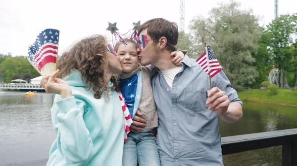4 Temmuz Bağımsızlık Günü 'nü kutlayan mutlu genç aile portresi. Park' a karşı milli bayrak sallayan ve kızını öpen. Ön görünüm