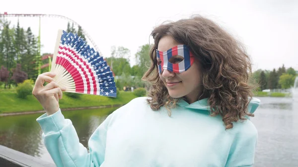 Çekici kıvırcık saçlı kadın, siluet ABD bayrağıyla ahşap yelpaze sallayarak ve Amerika Birleşik Devletleri parkında 4 Temmuz kutlamalarıyla mutlu..