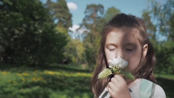 Close-up portret van bruine hazelaar meisje ruikt de witte appelboom bloemen met, glimlacht en kijkt naar de camera. Gelukkige kindertijd in tuin of bos tijdens de lente, genieten van de frisse natuur — Stockvideo