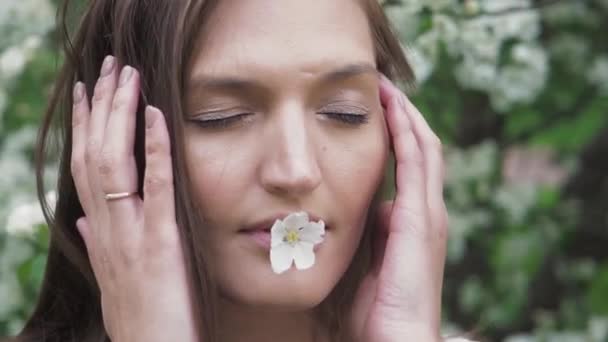 Κοντινό πλάνο πορτρέτο της ελκυστικής νεαρής γυναίκας με κλειστά μάτια που κρατά το μικρό λευκό λουλούδι στα χείλη και αγγίζοντας το πρόσωπο με τα χέρια στον ανθισμένο κήπο μηλιάς. Ομορφιά, υγεία, φρεσκάδα — Αρχείο Βίντεο