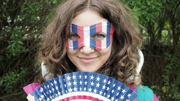 Karnaval maskeli mutlu kıvırcık saçlı kadın ve siluetli ahşap yelpaze Amerikan bayrağıyla Amerika Birleşik Devletleri parkında 4 Temmuz kutlamalarında gülümsüyor..