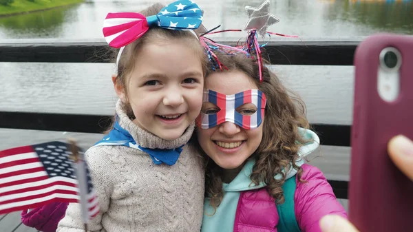 Sevimli küçük bir kız ve kadın Amerikan bayrağı takıyor, video konferansında akıllı telefondan konuşuyor, 4 Temmuz 'u kutlayan kameraya gülümsüyor. Teknoloji yaşam tarzı mobil kullanım kavramı