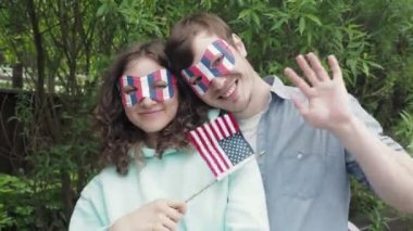 Komik maskeli mutlu vatansever genç çift ve küçük vatansever bayrak ulusal beyaz-mavi-kırmızı renkler Amerikan bayramında eğlence parkında 4 Temmuz 'da bağımsızlığı kutlarken kameraya bakın.