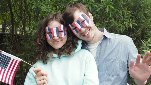 Komik maskeli mutlu vatansever genç çift ve küçük vatansever bayrak ulusal beyaz-mavi-kırmızı renkler Amerikan bayramında eğlence parkında 4 Temmuz 'da bağımsızlığı kutlarken kameraya bakın.