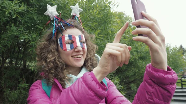 Cute happy curly hair woman in funny mask and silly carnival horns with silhouette usa national flag making video call with family and friends using phone, smiles enjoying 4th of July USA celebration